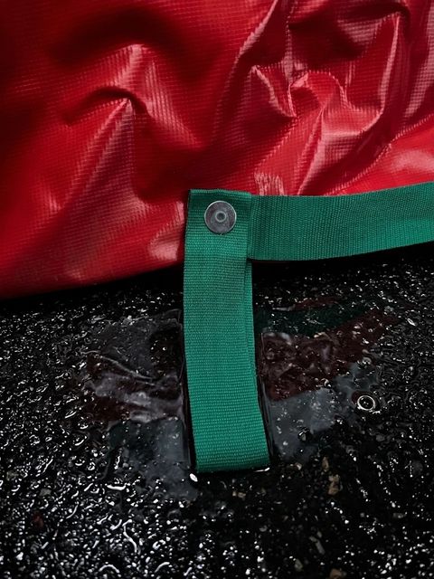 Close-up of a green webbing carry loop riveted to the red vinyl bag, on a wet driveway.